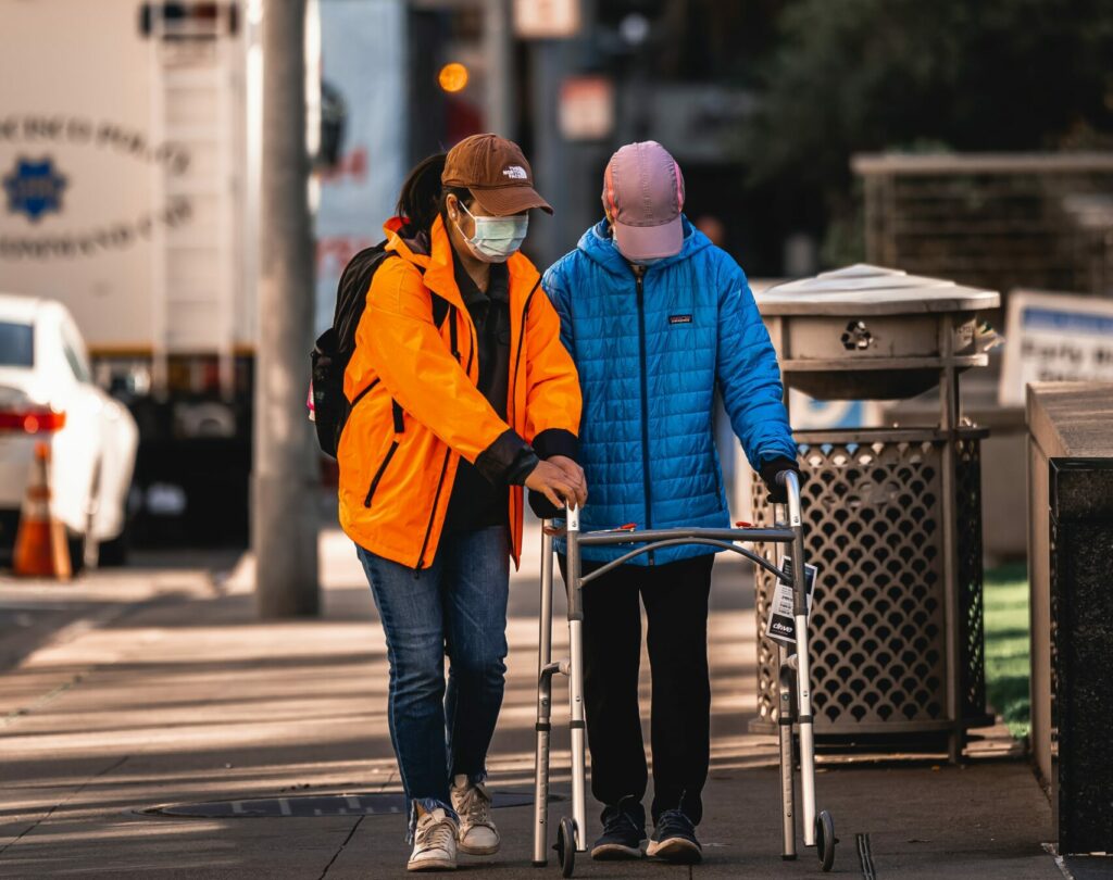 A person wearing a mask helping another person walk down the sidewalk with a walker.