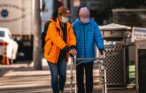 A person wearing a mask helping another person walk down the sidewalk with a walker.