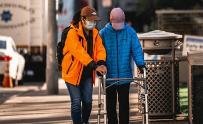 A person wearing a mask helping another person walk down the sidewalk with a walker.